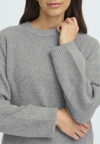 Szürke kötött pulóver bordás textúrával, széles mandzsettákkal és kerek nyakkal, egy karjaikat keresztbe tett személyen.