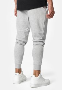 Person står i lysegrå joggerbukser og hvide sneakers, med hænderne afslappet langs siden, vist fra ryggen mod en hvid baggrund.