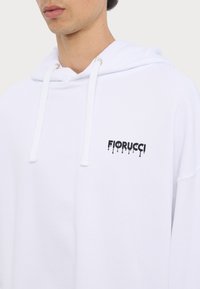 Hvit hettegenser med snøring, med en svart "Fiorucci"-logo på brystet. Laget av mykt stoff; avslappet passform.