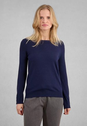 Donna bionda che indossa un maglione a maniche lunghe blu navy e pantaloni grigi, in piedi di fronte a uno sfondo grigio uniforme.