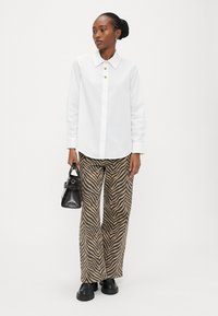 Kvinne iført hvit skjorte med knapper, brune og svarte zebra-mønstrede vide bukser, svarte sko, holder en svart håndveske, står på hvit bakgrunn.