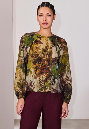 Frau mit geflochtenem Haar, die eine langärmelige, transparente Bluse mit grünen und braunen Blattmustern und dunkelroter Hose trägt, steht vor einer schlichten Wand.