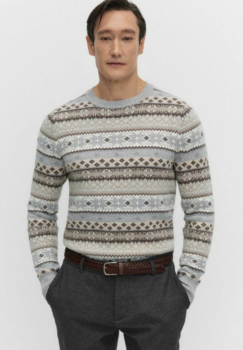 Man met kort donker haar, gekleed in een grijs, wit en bruin geblokt truitje, donkergrijze broek en een bruin gevlochten riem, met de handen in zijn zakken.