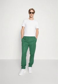 Calças desportivas verdes com cintura elástica, riscas laterais brancas e uma t-shirt branca de manga curta. Usadas com óculos de sol e sapatilhas brancas.