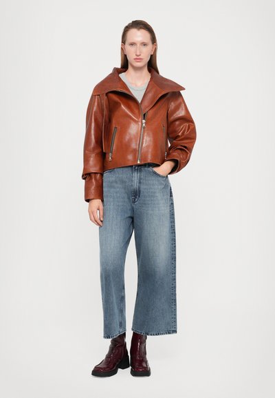 Giacca cropped in pelle marrone con un ampio colletto, abbinata a pantaloni in denim a gamba larga blu chiaro e stivaletti alla caviglia borgogna.
