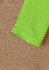 Brunt stickat tyg övergår till en grön stickad ärm. Ärmen har en ribbad manschett, och båda tygerna har en slät yta.