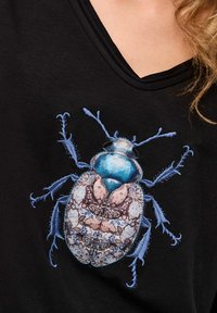 Close-up of a black shirt with a detailed embroidered blue beetle featuring intricate patterned wings on a person’s chest.