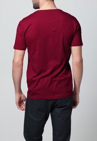 Burgundské bavlněné tričko s krátkým rukávem a kulatým výstřihem, z pohledu zezadu, v kombinaci s tmavě modrými džíny. Jednoduchý design v jedné barvě.
