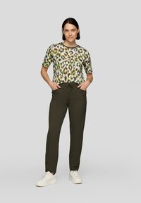 Frau steht mit Händen in den Taschen und trägt ein kurzärmliges Leopardenmuster-Oberteil, dunkelgrüne Jogginghosen mit Kordelzug und weiße Turnschuhe vor einem grauen Hintergrund.