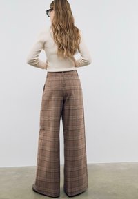 Pantalon taille haute, coupe large en tissu écossais marron, avec un motif à carreaux et des poches latérales, associé à un haut ajusté beige clair.