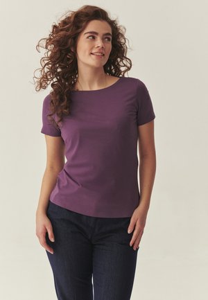 Młoda kobieta o kręconych włosach, ubrana w fioletową bluzkę z krótkim rękawem i ciemne spodnie, uśmiecha się i patrzy w bok.