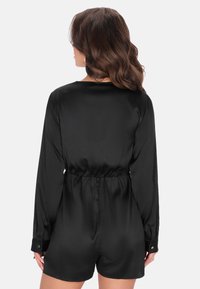 Černý saténový overal s dlouhými rukávy, elastickým pasem a knoflíkovými manžetami; hladká textura a přiléhavý střih zvýrazňují celkovou siluetu.