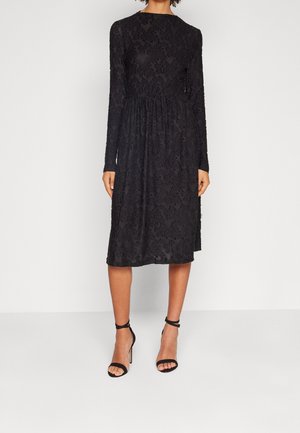 Woman wearing a black long-sleeve knee-length dress with textured floral patterns and black ankle-strap high-heeled sandals.