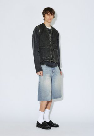Ung person står iført en falmet sort langærmet skjorte med sort ærmeløs vest, lyse denim knælange shorts, hvide sokker og sorte sko.