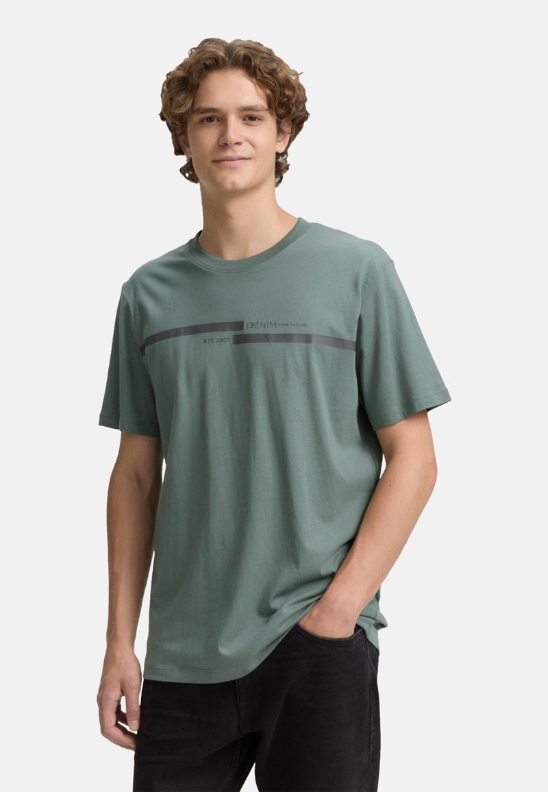 Grøn bomulds t-shirt med rund hals, der har et horisontalt sort stribedesign og trykt tekst, brugt sammen med sorte bukser.