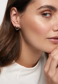 Gul hjerteformet studørering med en glat, poleret finish. Modellen har langt, brunt hår, der står i kontrast til smykkernes glans.