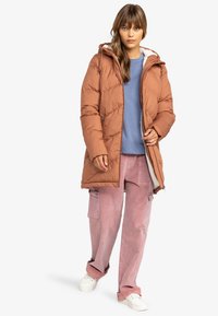 Bruine gewatteerde jas met een capuchon, met een gequilte uitstraling en een zachte binnenkant. Gecombineerd met lichtroze corduroy broek en witte sneakers.