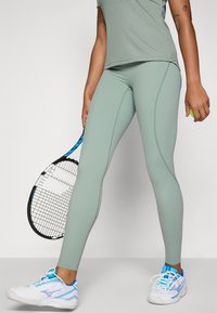 Leggings leggeri verdi in materiale traspirante, con design aderente e dettagli delle cuciture, abbinati a scarpe da ginnastica bianche e blu. In mano tiene una racchetta da tennis.