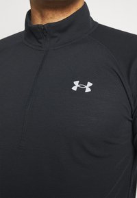Černý pulovr se čtvrtým zipem vyrobený z texturované látky, s výrazným bílým logem Under Armour na hrudi.