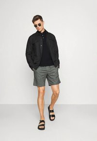 Μαύρο σακάκι με κουμπιά πάνω από μαύρο πόλο πουκάμισο, συνδυασμένο με γκρι σορτς και μαύρα σανδάλια. Σημαντικές λεπτομέρειες περιλαμβάνουν γυαλιά ηλίου και μια χαλαρή στάση.