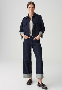 Giacca di denim scuro con due tasche sul petto, indossata sopra una maglietta bianca. Abbinata a pantaloni ampi in denim scuro a vita alta con orli ripiegati.