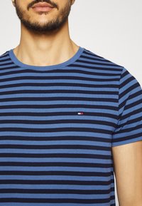 Modro-navy pruhované tričko s kulatým výstřihem. Obsahuje malé logo na levé straně hrudi. Vyrobeno z měkké a pohodlné látky.