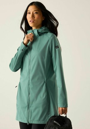 Vrouw draagt een tealkleurige capuchonvest met rits en houdt een zwarte Regatta Great Outdoors rugzak vast tegen een effen achtergrond.