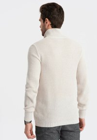 Pull en tricot couleur crème avec un col montant, présentant un motif texturé et des côtes aux poignets et à l'ourlet.