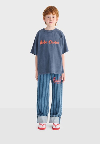 Enfant aux cheveux roux debout, portant un T-shirt bleu marine surdimensionné avec le texte orange "Bobo Choses", un jean rayé bleu avec des revers, et des sandales rouges.