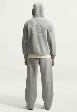 Person trägt einen grauen Nike Sportswear Hoodie und passende Jogginghose und steht mit dem Rücken zu sehen vor einem einfarbigen hellen Hintergrund.