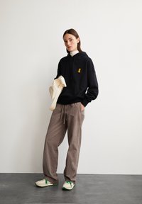 Haut à capuche noir avec un logo en ruban jaune, associé à un pantalon en velours côtelé marron et des baskets multicolores. Le modèle tient un sac beige.