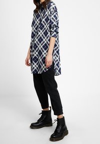 Camicia a quadri nei colori blu navy, bianco e viola; si abbina a pantaloni cropped neri e stivali neri con lacci; vestibilità rilassata, maniche lunghe.