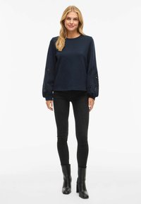 Donna che indossa un top lungo a maniche navy con modelli floreali ritagliati sulle maniche, jeans skinny neri e stivali neri con tacco alla caviglia, in piedi.
