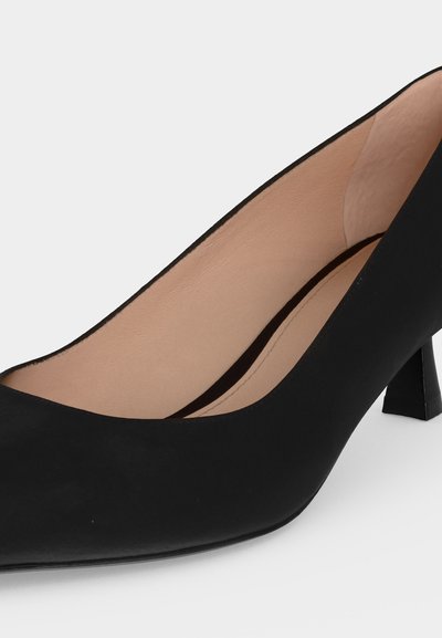 Escarpin noir à bout fermé et talon bas, intérieur en cuir beige et talon carré légèrement incurvé sur un fond clair.
