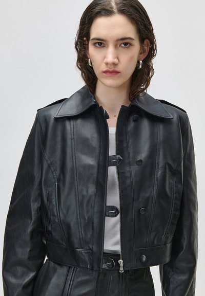 Νεαρή γυναίκα με κοντά, σγουρά μαλλιά φοράει ασημένια κρίκους σκουλαρίκια, μαύρο δερμάτινο μπουφάν πάνω από λευκή μπλούζα, αντιμετωπίζει προς τα εμπρός με ουδέτερη έκφραση.