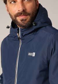 Veste imperméable bleu marine avec capuche, dotée d'une fermeture éclair à l'avant et d'un écusson logo. Texture lisse et cordons de serrage réglables sur la capuche.