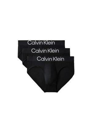 Homme debout pieds nus sur fond gris, portant un slip noir Calvin Klein avec ceinture à logo, mains derrière le dos.