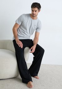T-shirt grigio a maniche corte con un piccolo logo, abbinato a pantaloni neri a vestibilità ampia. Persona seduta su un divano di colore chiaro e dalla forma arrotondata.