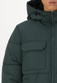 Green puffer jacket with a hood, features a front zipper, adjustable drawstring, and a large chest pocket with flap detailing.