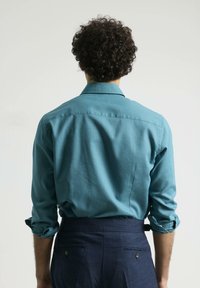 Camisa de manga larga turquesa con cuello clásico y puños doblados, combinada con pantalones oscuros de color marino que tienen bolsillos traseros y detalles de botones.