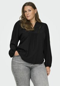 Blusa nera a maniche lunghe con scollo a V, caratterizzata da pieghe sottili e un motivo testurizzato. Abbinata a jeans grigi aderenti.