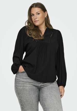 Blouse noire à manches longues avec un col en V, présentant des plis subtils et un motif texturé. Associée à un jean gris ajusté.