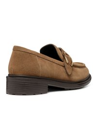 Mocasines de ante marrón con una solapa decorativa y detalle de hebilla metálica. Cuenta con una suela de goma marrón texturizada y baja. Textura suave.