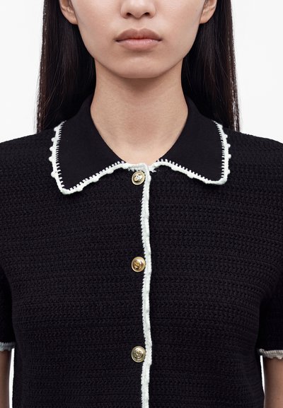 Femme portant un haut en maille noire avec un col bordé de blanc et des boutons dorés sur le devant, avec de longs cheveux foncés visibles.