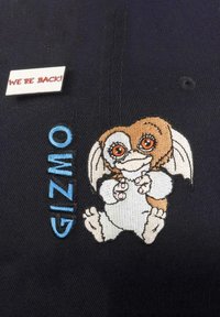 Der schwarze Stoff zeigt eine gestickte Figur mit großen Augen und braunem Fell. Eine weiße Anstecknadel trägt die Aufschrift "WIR SIND ZURÜCK!" in roter Schrift.