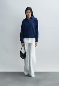 Marineblauer Pullover mit Viertel-Reißverschluss und rotem Logo, kombiniert mit weißen Hosen in weiter Beinweite und einer kleinen schwarzen Handtasche. Gleichmäßige Textur durchgehend.