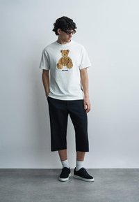 Jeune homme portant des lunettes de soleil, un t-shirt blanc avec un motif ours en peluche, un pantalon court noir, des chaussettes blanches et des baskets noires, debout de manière décontractée.