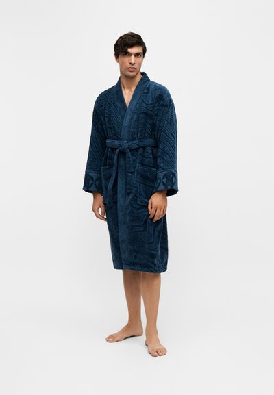 Jeune homme aux cheveux foncés portant un peignoir bleu foncé texturé, noué à la taille, debout pieds nus devant un fond clair uni.