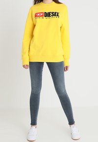 Sudadera amarilla con el logo negro de "DIESEL" y el texto "DENIM DIVISION", emparejada con jeans ajustados grises y zapatillas blancas.