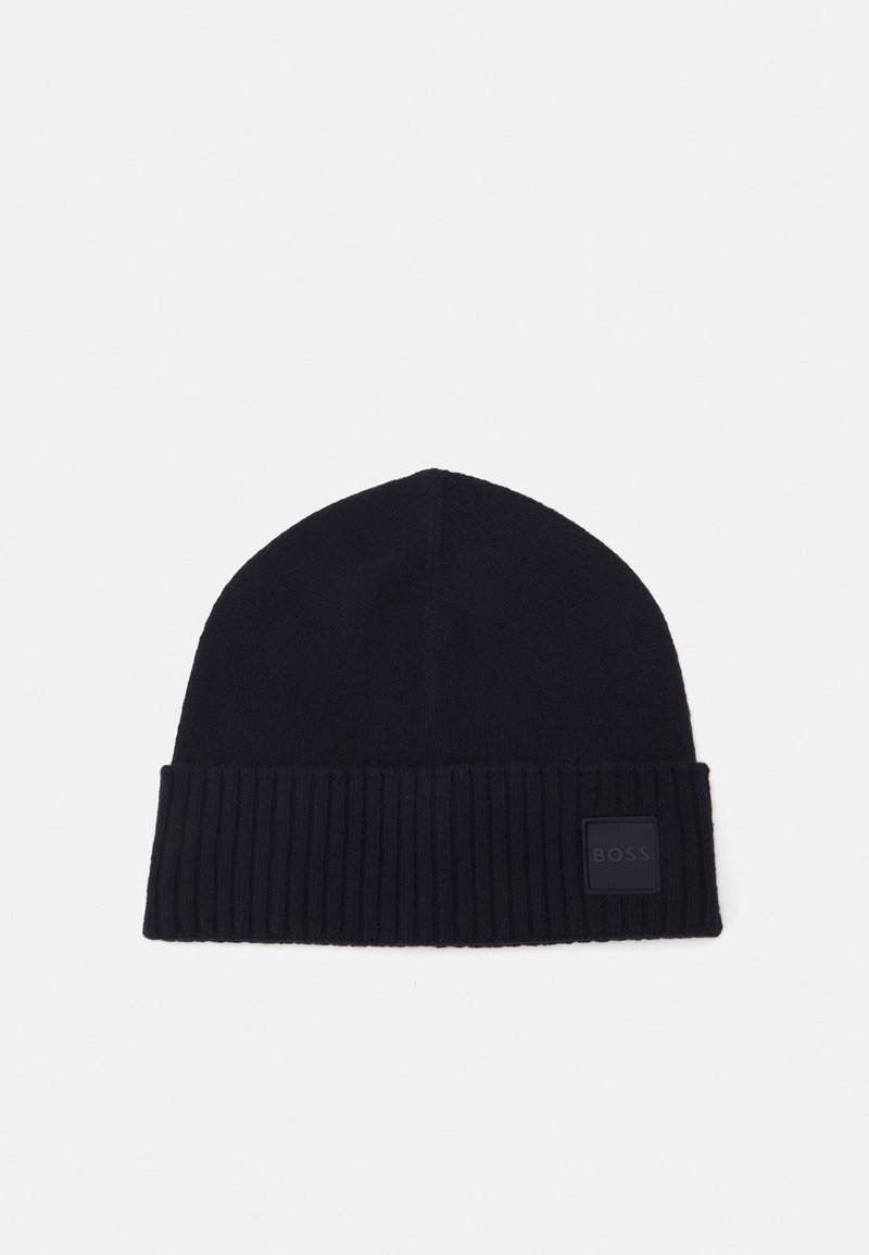 Strikket marineblå lue med ribbestrikket bånd og et kvadratisk logomerke. Myk tekstur, tettsittende design som smalner mot toppen.